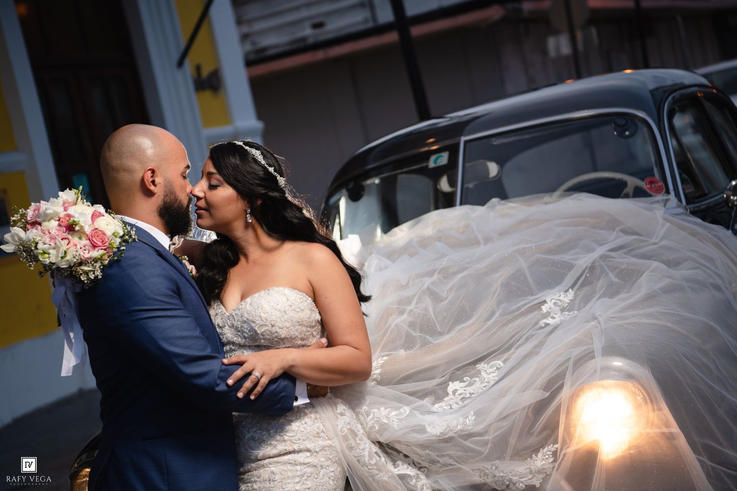 Bride and groom share a romantic kiss near a vintage black car, with her veil flowing in the breeze.