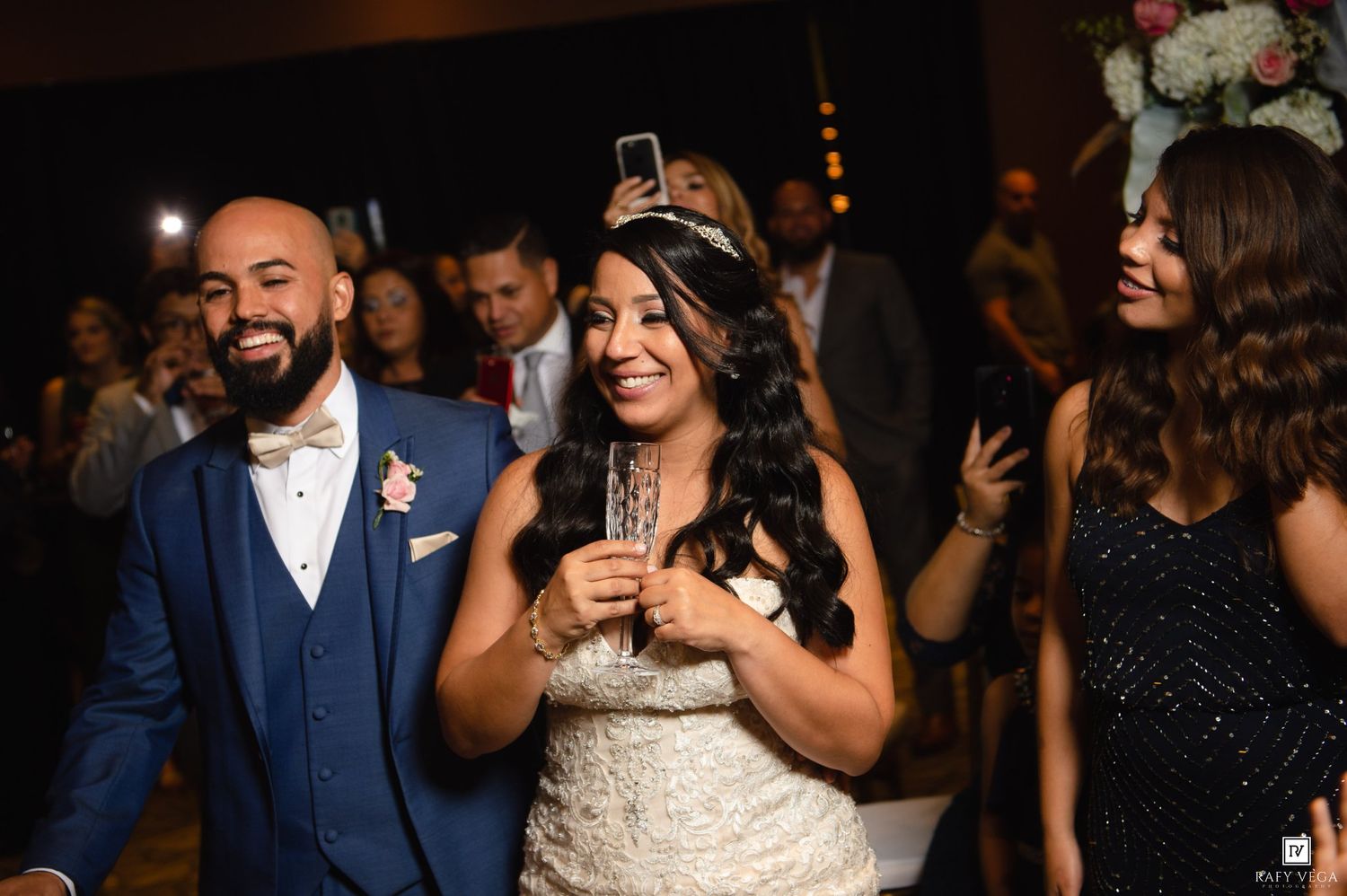 Bride in tiara and groom in navy suit smiling joyfully at wedding reception with guests dancing in background.