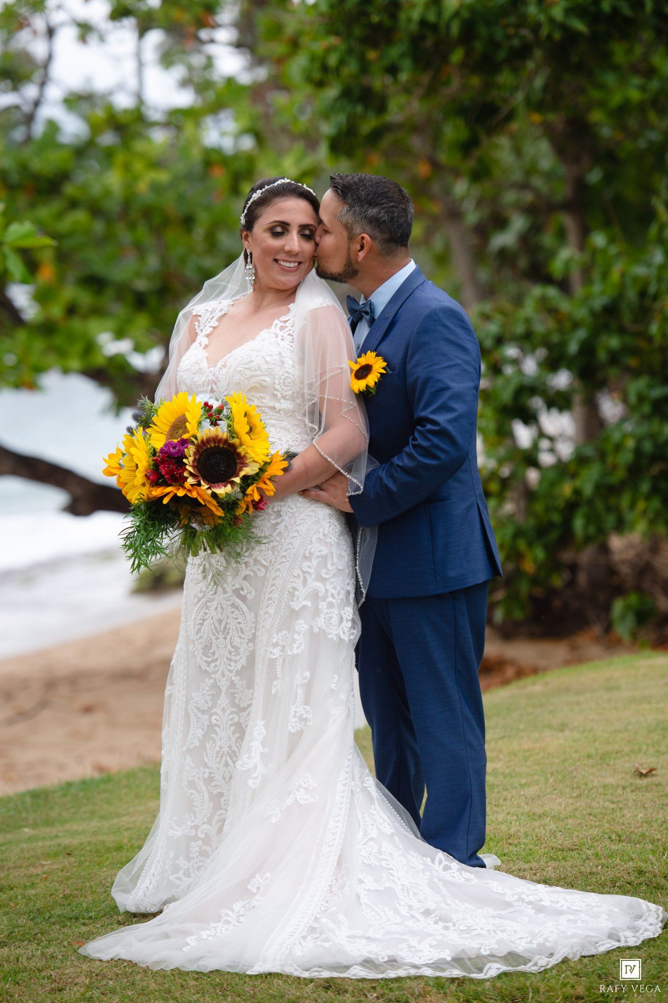 Bride in lace gown holding sunflower bouquet receives kiss from groom in blue suit at beach wedding.