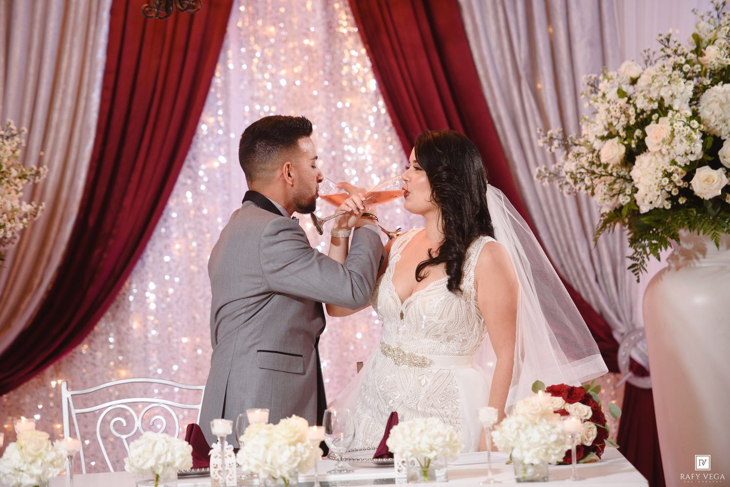 Bride and groom sharing a champagne toast at their elegant wedding reception with red curtains and floral decor.