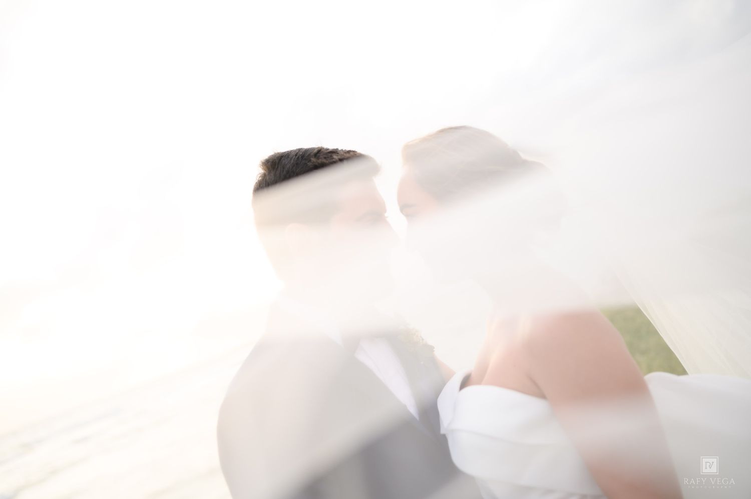 Overexposed romantic wedding photo of couple nearly kissing, draped in flowing white veil in bright light.