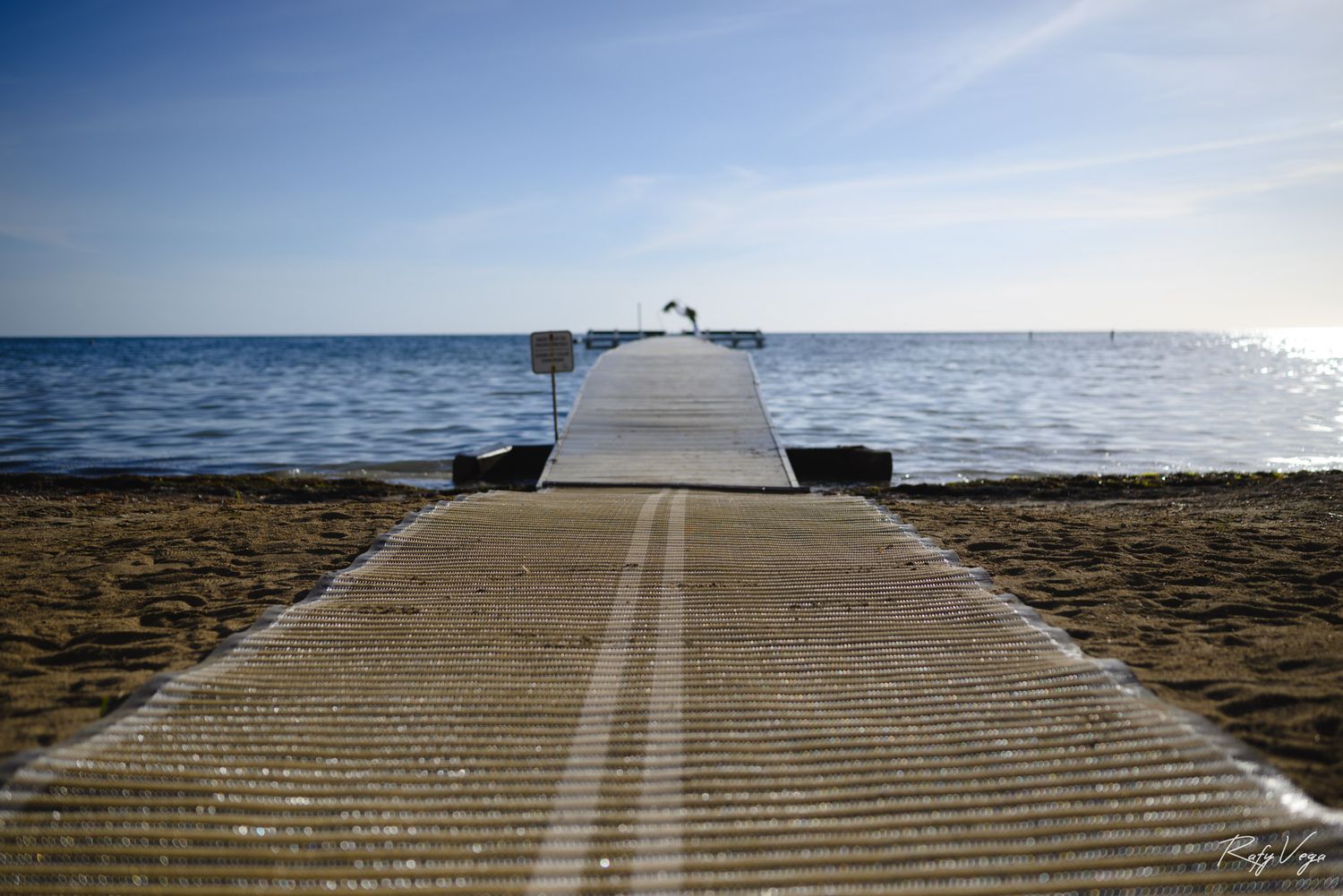 A wooden pier extending into calm blue ocean waters on a sandy beach under a partly cloudy sky.