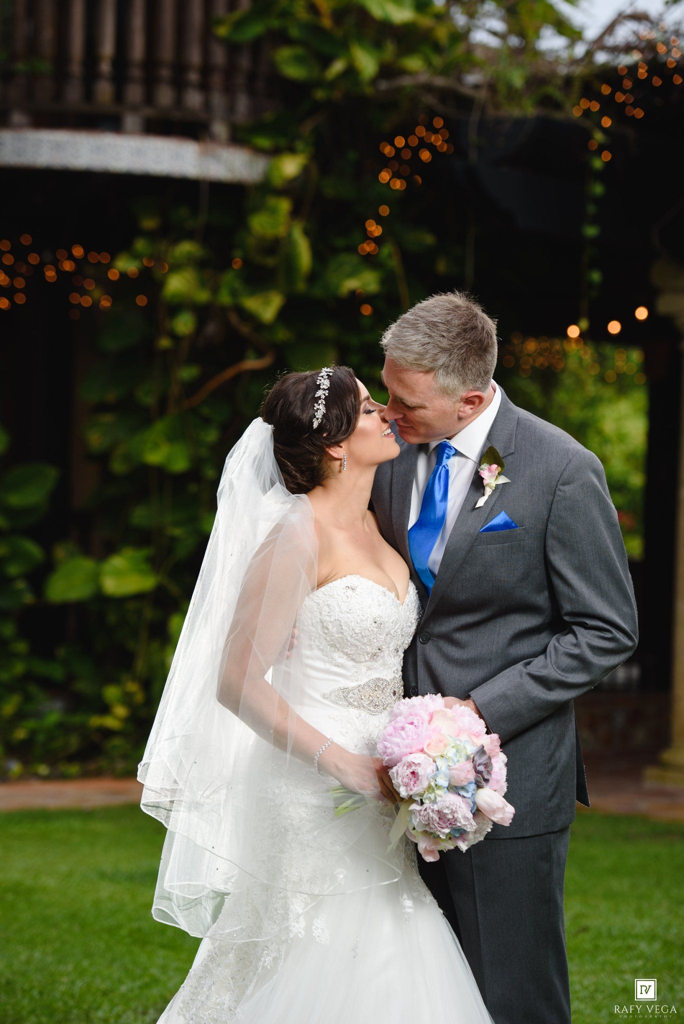 Bride and groom share a romantic moment outdoors, bride holding pink bouquet in white gown with veil, groom in gray suit.