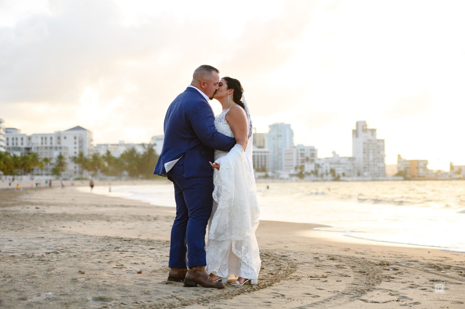 Couple sharing a romantic beach kiss at sunset, groom in blue suit and bride in white dress with city skyline backdrop.