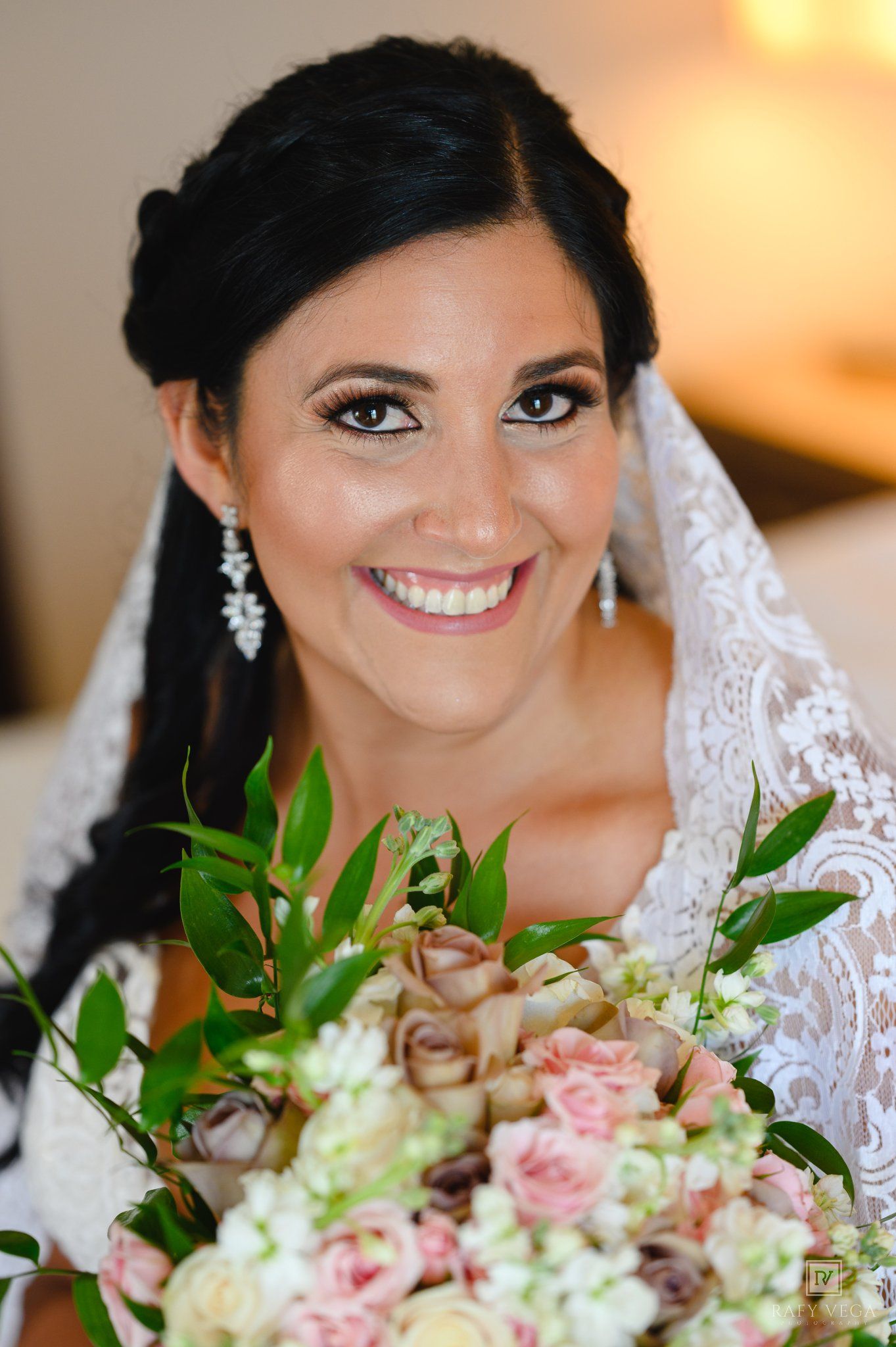 Smiling bride holding a bouquet of pink and white roses, wearing a lace veil and elegant drop earrings.