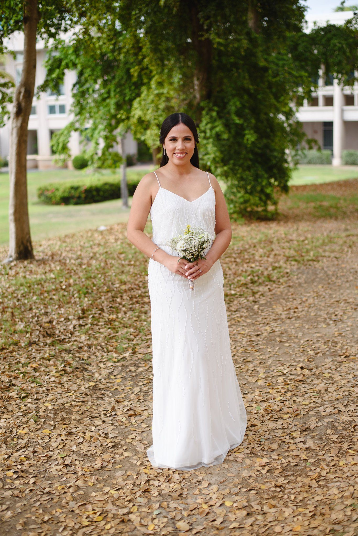 Bride in white spaghetti strap gown holding small bouquet of white flowers, standing on leaf-covered path in park.