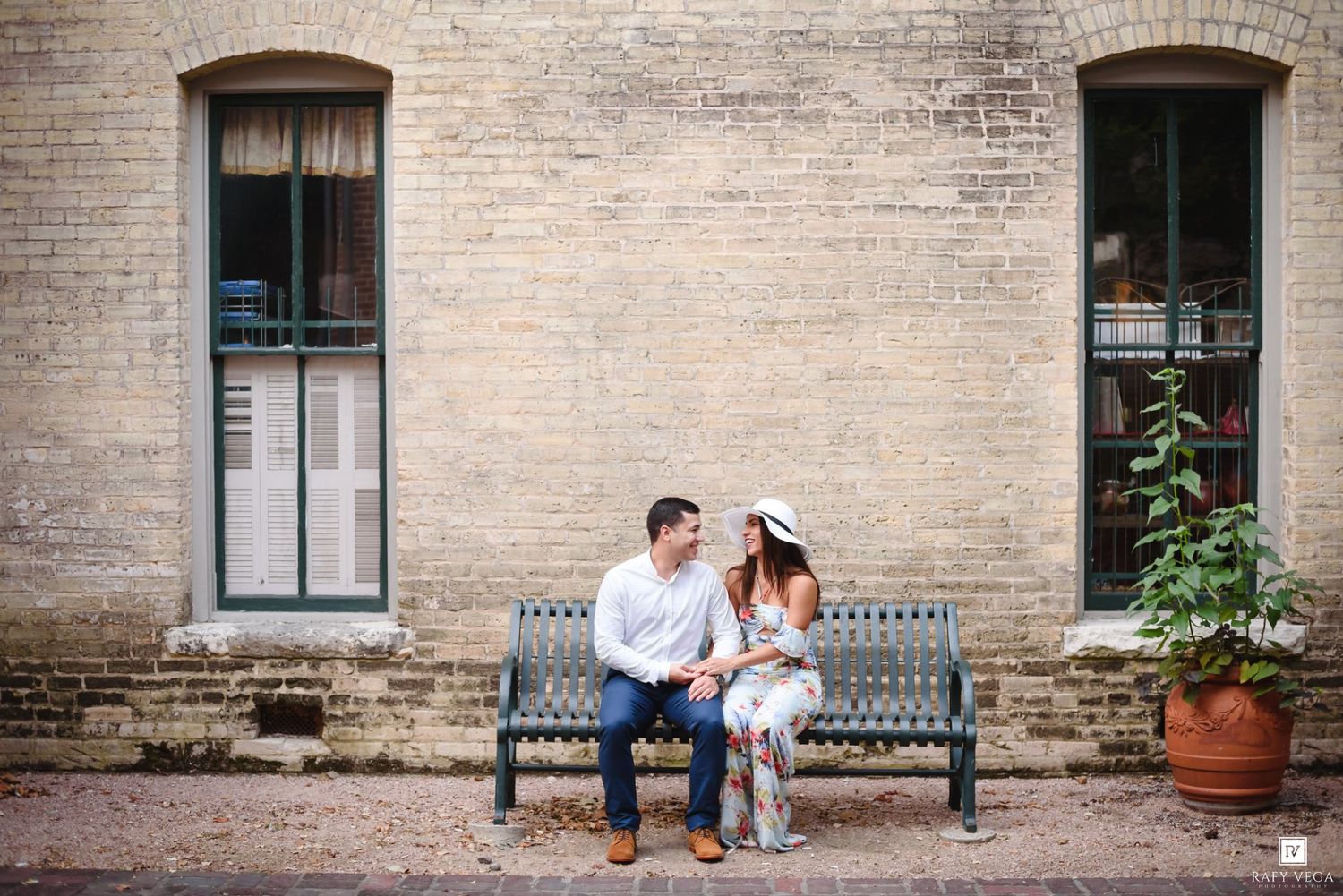 Couple sitting on a bench smiling at each other against a rustic brick wall with large windows.