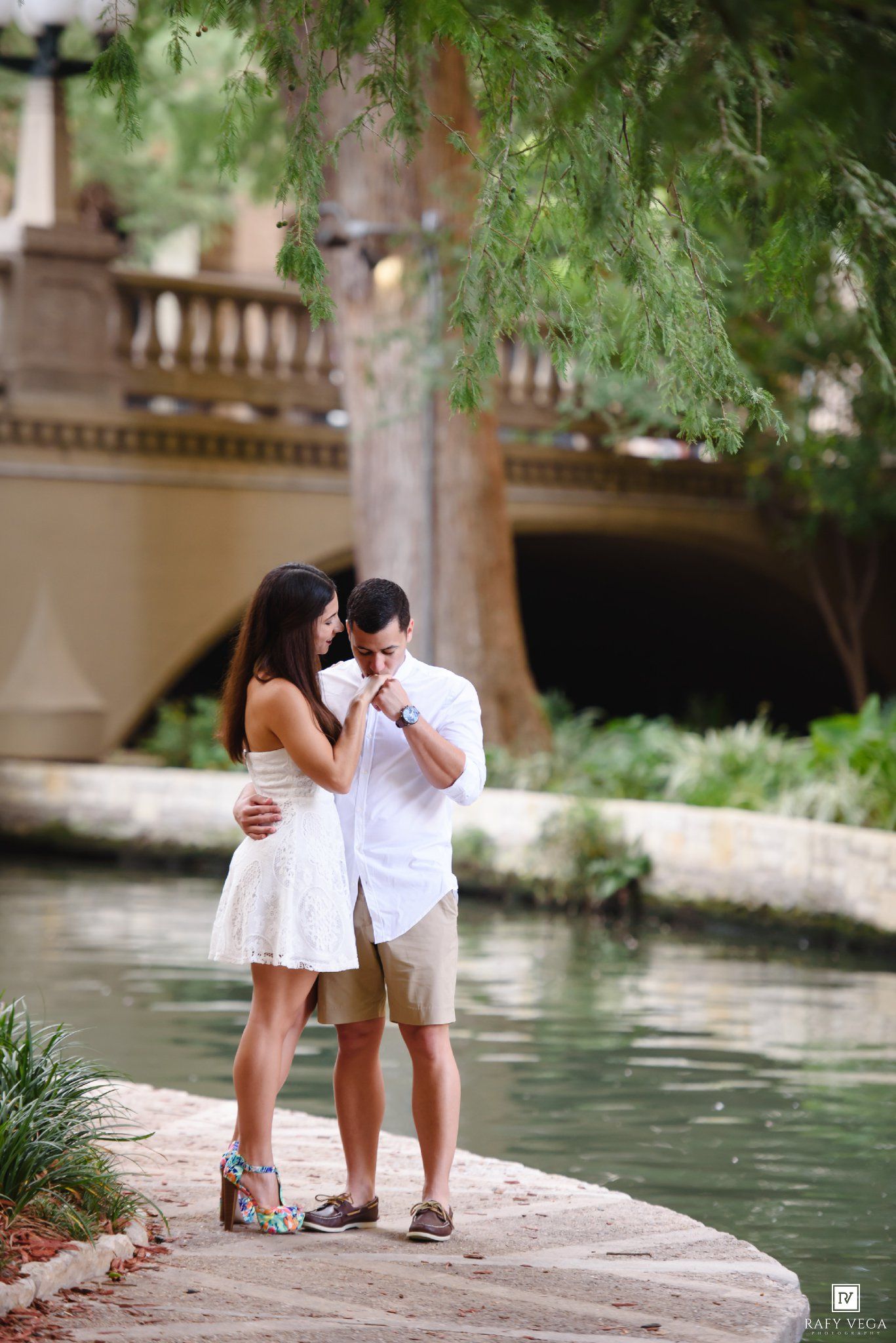Romantic couple standing by a river on the San Antonio Riverwalk, man kissing woman's hand near a stone bridge.