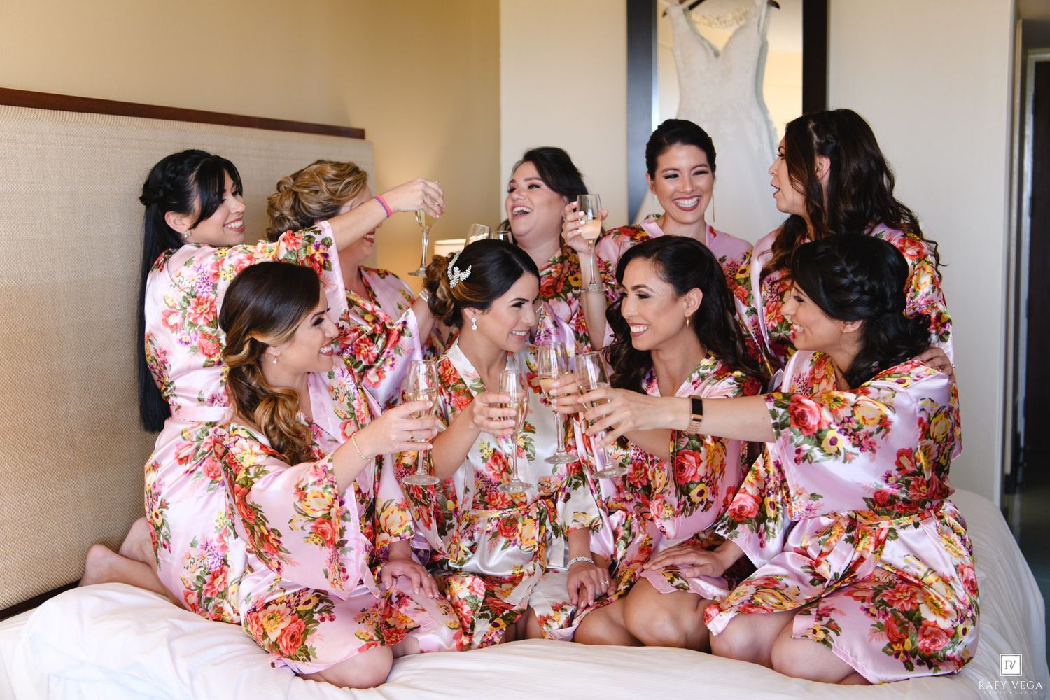 Bridesmaids in matching floral robes toast champagne together on a bed before the wedding.