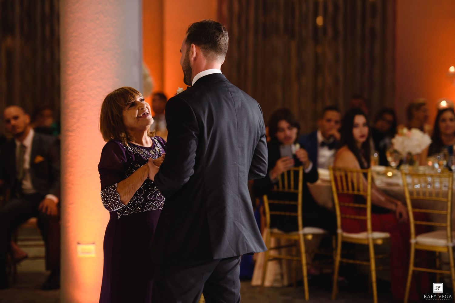 Mother and son share an emotional dance at a wedding reception with warm amber lighting and seated guests.
