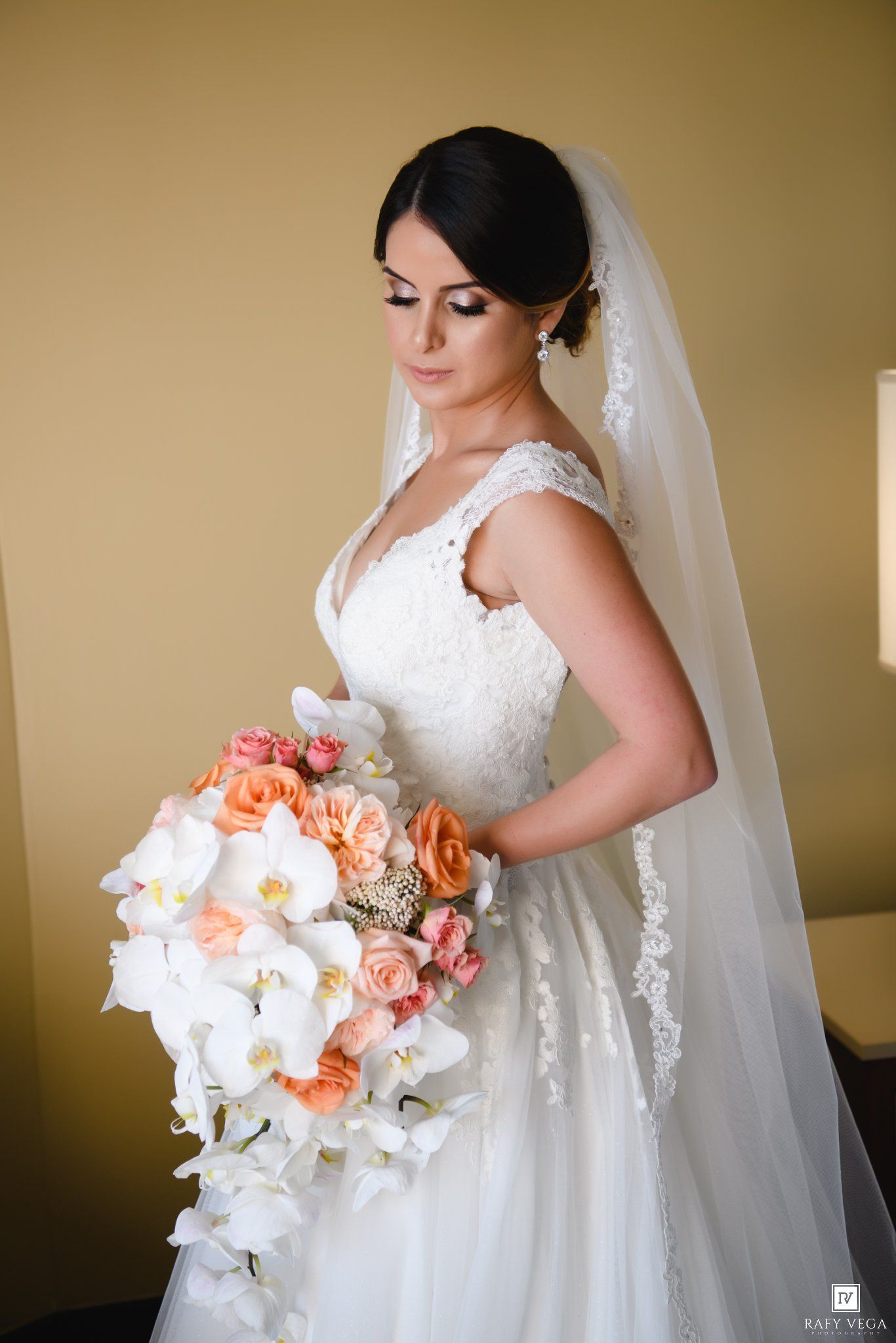 Bride in white lace gown and veil holds cascading bouquet of white orchids and peach roses.