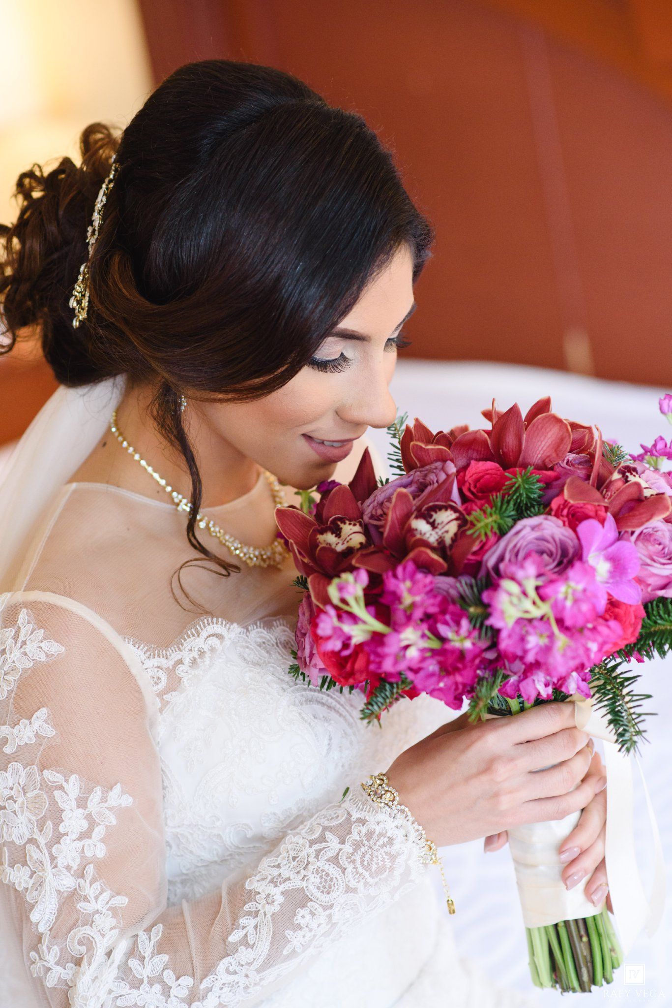 Bride in lace gown holding vibrant pink and red wedding bouquet, looking down with elegant updo hairstyle.