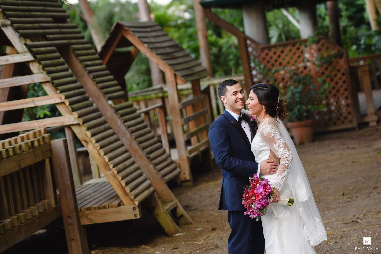 A bride and groom share a romantic moment outdoors near wooden staircases, she holds a vibrant pink bouquet.