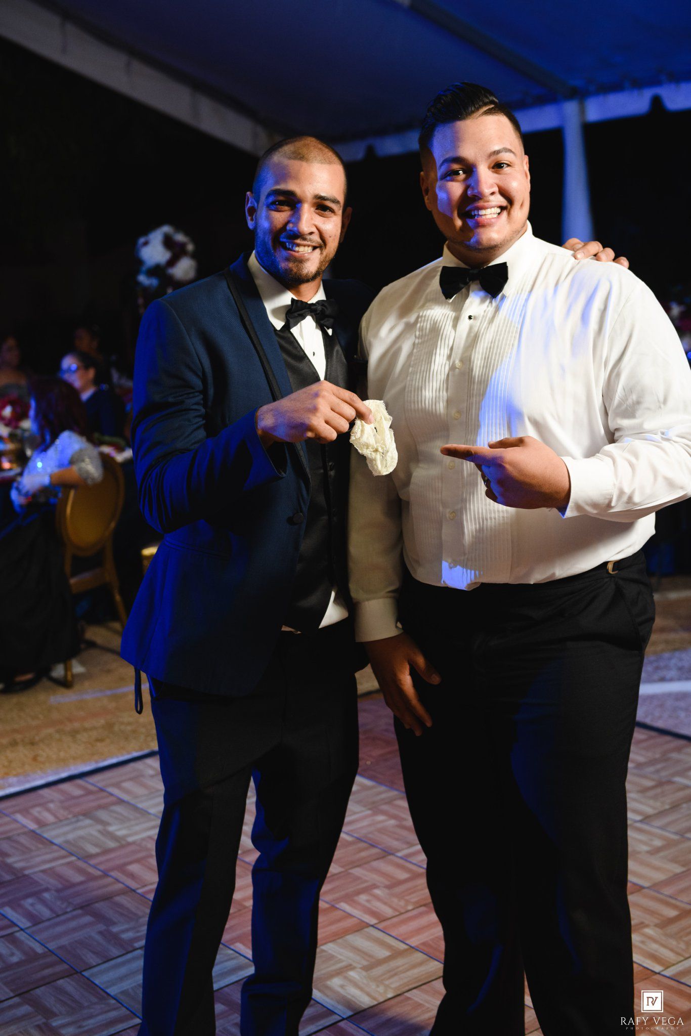 Two men in tuxedos smile and pose together at a formal wedding reception event.