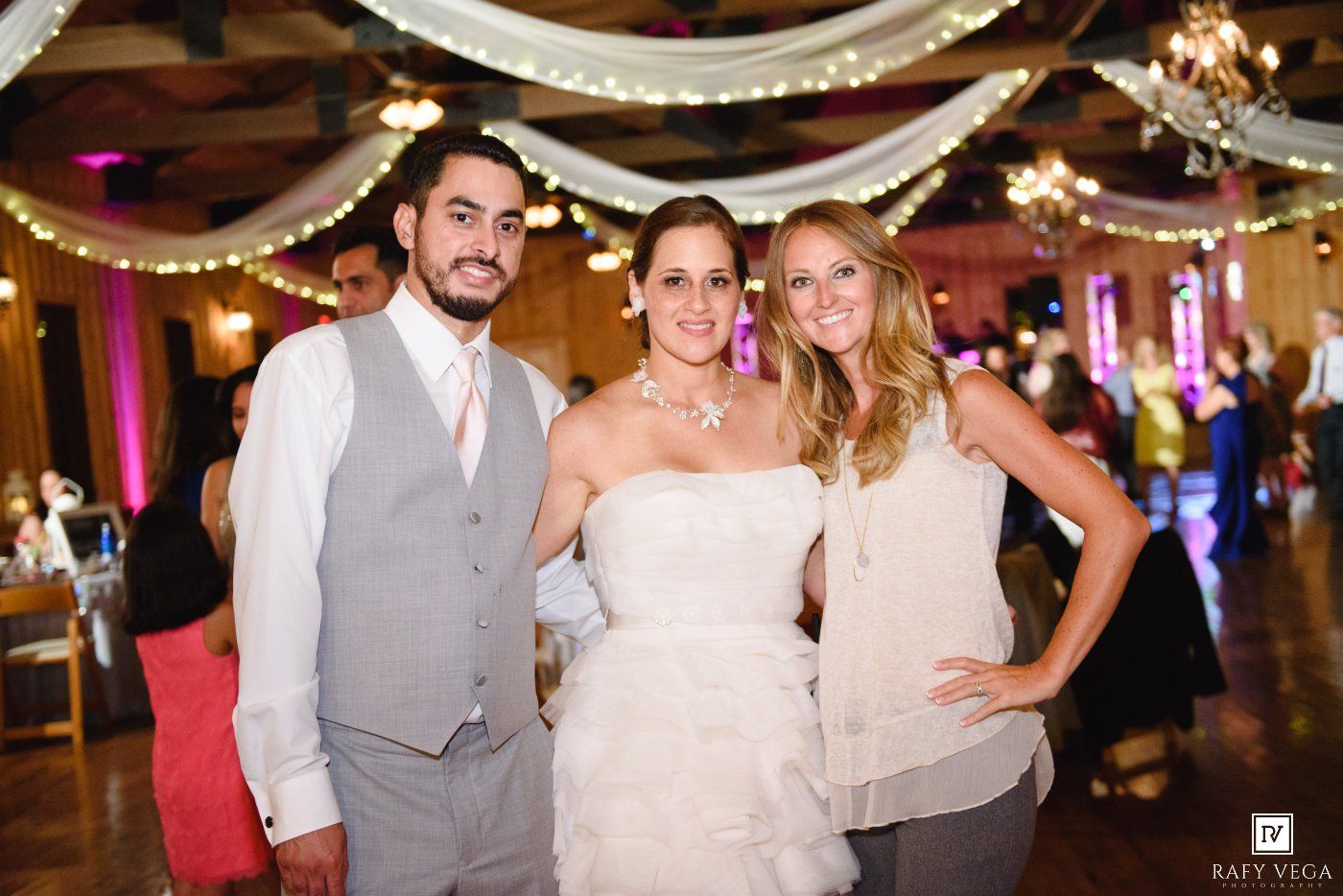 Bride in white strapless gown poses with groom in gray vest and woman in white dress at elegant wedding reception.