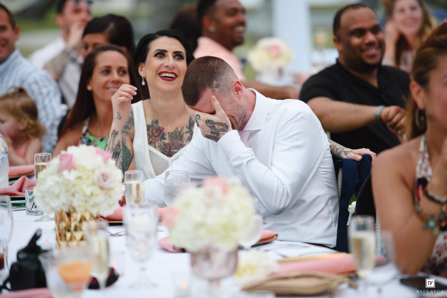 Couple laughing at outdoor wedding reception dinner table with floral centerpieces and guests in background.