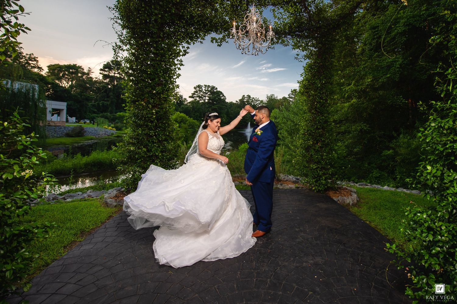 Bride and groom dancing on garden path under chandelier at sunset, surrounded by lush greenery.