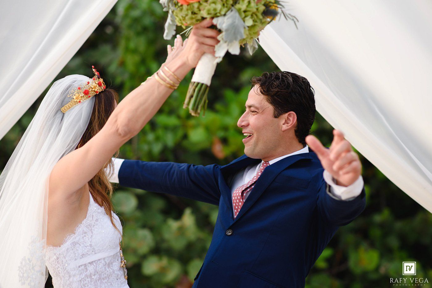 Joyful bride and groom celebrate at outdoor wedding ceremony, groom in navy suit raising bouquet under white draped arch.
