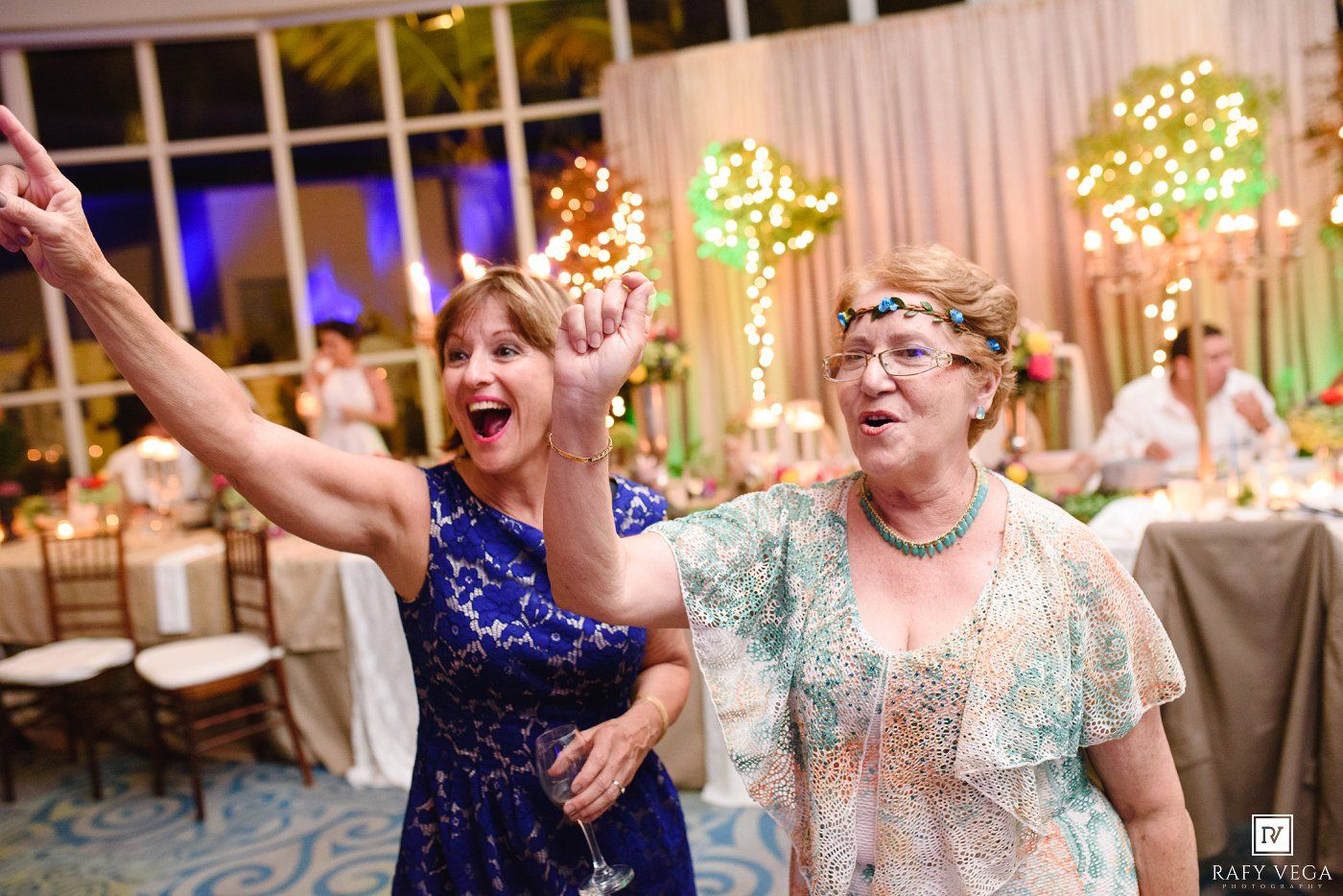Two women dancing joyfully at an elegant wedding reception with colorful floral decorations.