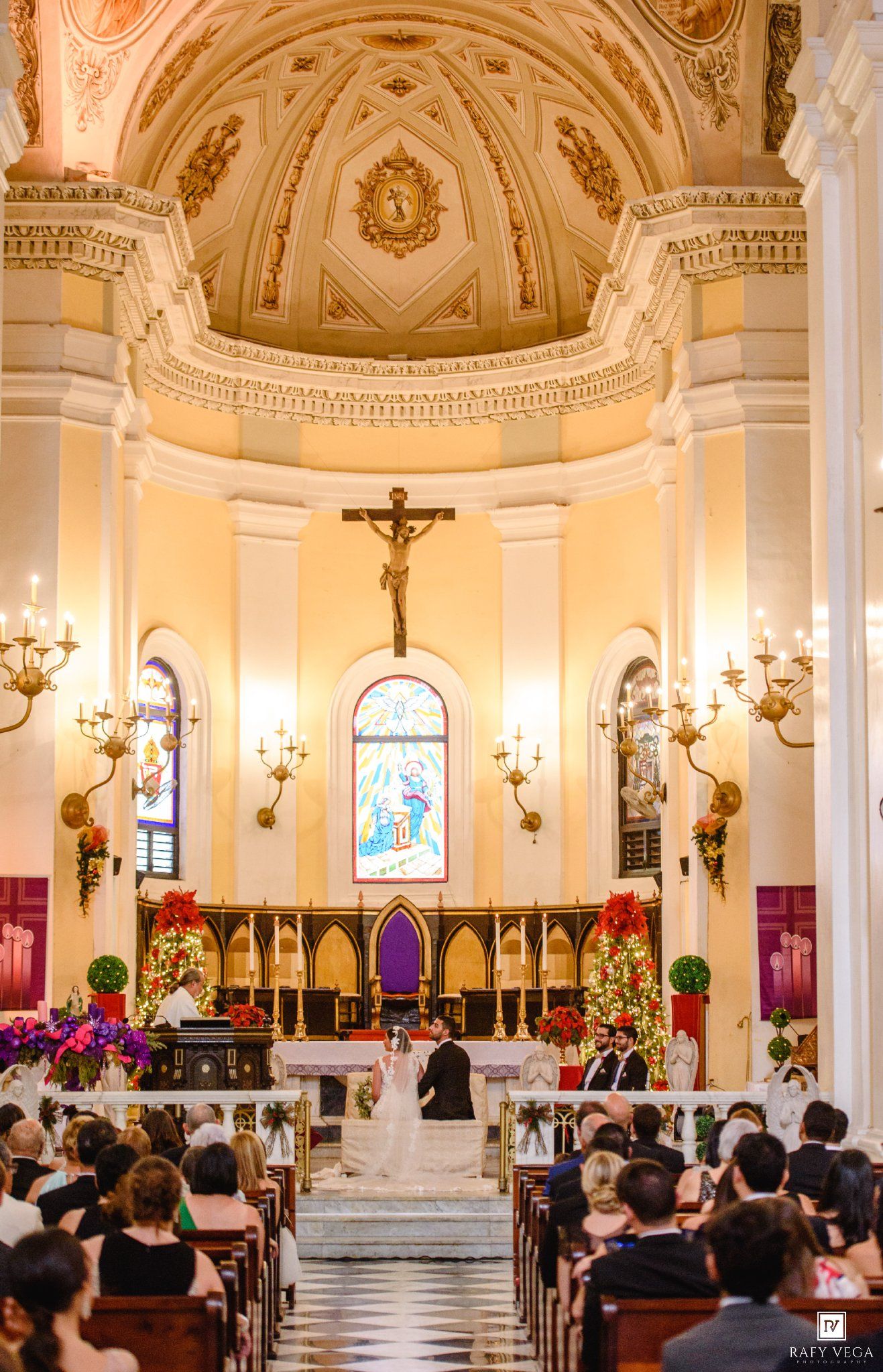 Elegant Catholic church wedding ceremony with ornate gold ceiling, stained glass windows, and guests seated in wooden pews.