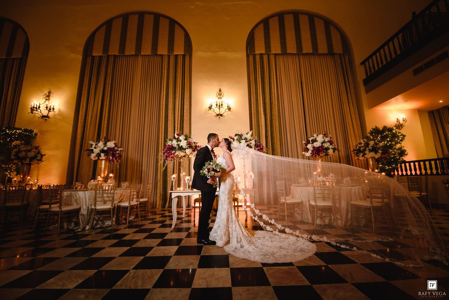 Bride and groom share a romantic kiss in an elegant ballroom with checkered floors and floral arrangements.