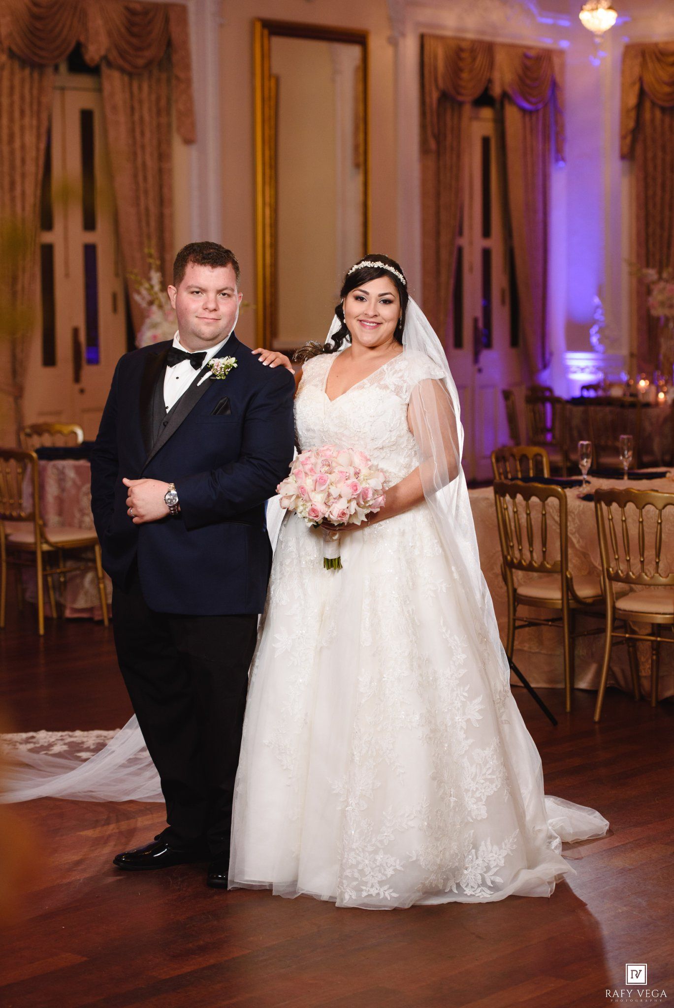 Bride in lace ball gown with veil and groom in navy tuxedo pose together in elegant wedding reception hall.