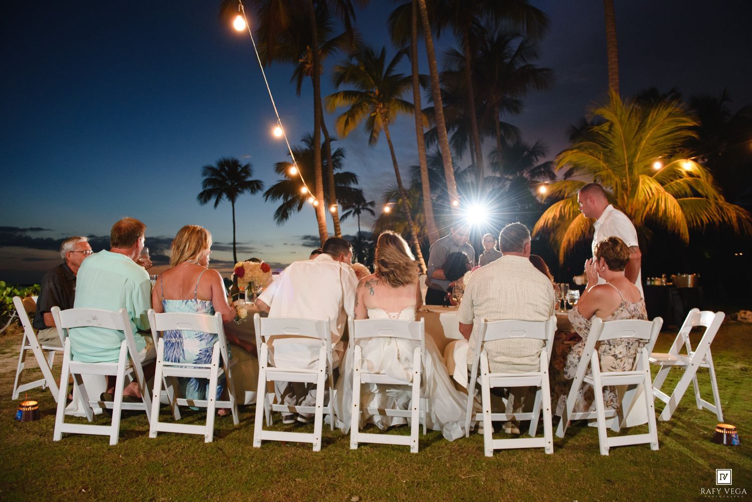Outdoor beach wedding reception at dusk with guests seated at tables under string lights surrounded by palm trees.