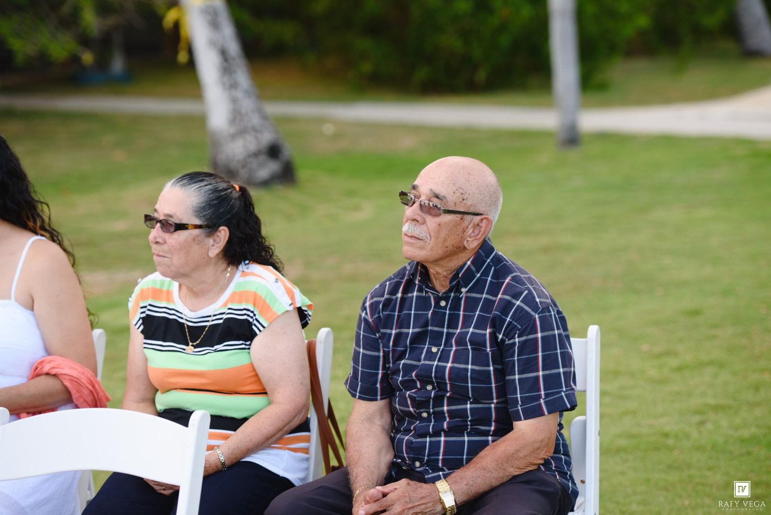 Two adults sitting on white chairs outdoors at a tropical event, with palm trees and green lawn in the background.