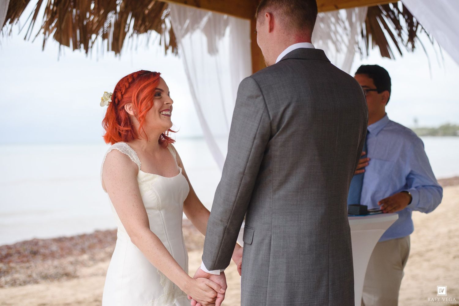 Bride with red hair and groom holding hands during beach wedding ceremony under thatched canopy with officiant.
