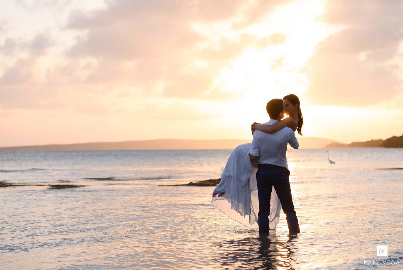 Couple embracing at sunset on a beach, man lifting woman in white dress with ocean waves and pastel sky.
