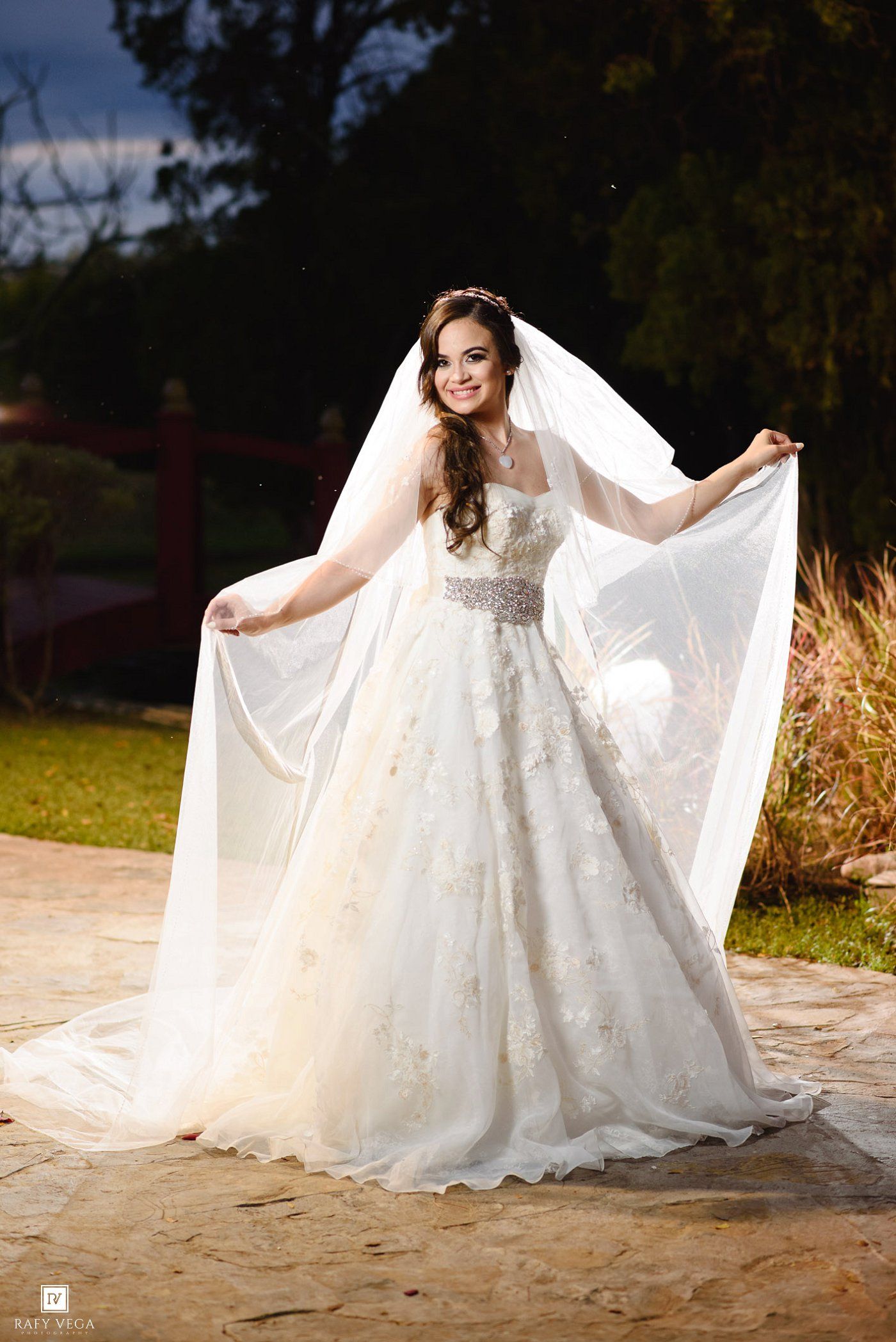 Bride in white floral ball gown with embellished belt and long veil spread wide, posing outdoors at dusk.