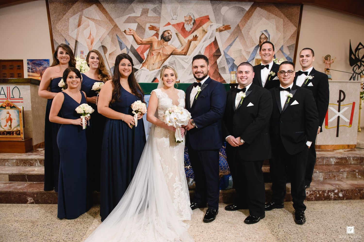 Wedding party poses in church with bridesmaids in navy gowns, groomsmen in tuxedos, and bride and groom in center.