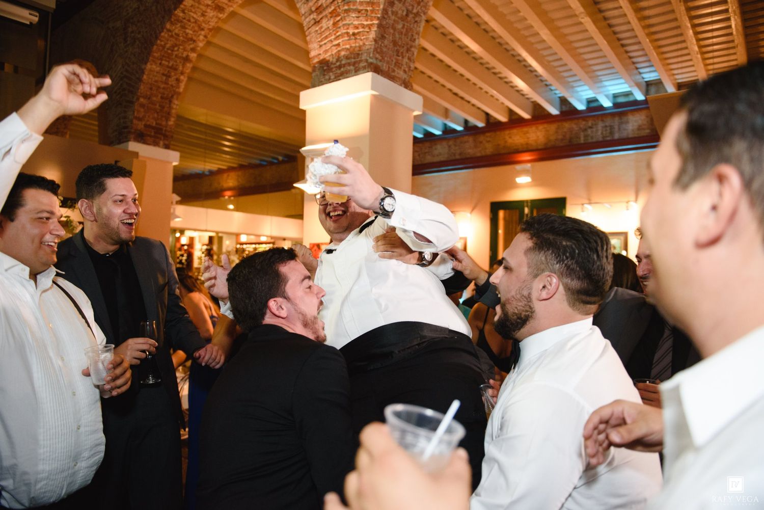 Groom lifted by groomsmen during lively wedding reception celebration indoors.
