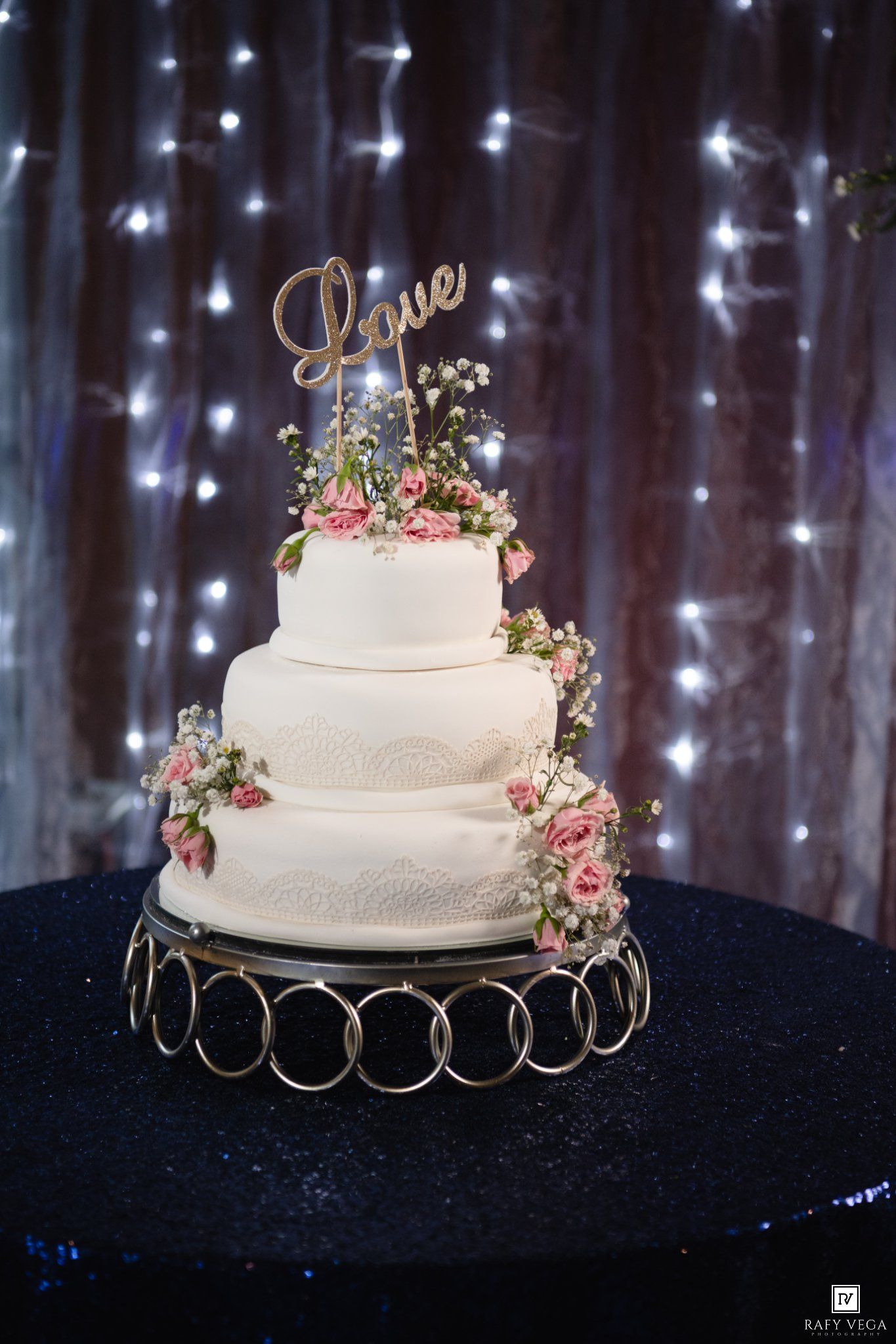 Elegant 4-tier white wedding cake with pink roses, lace details, and a gold Love topper on a ring stand with fairy lights.