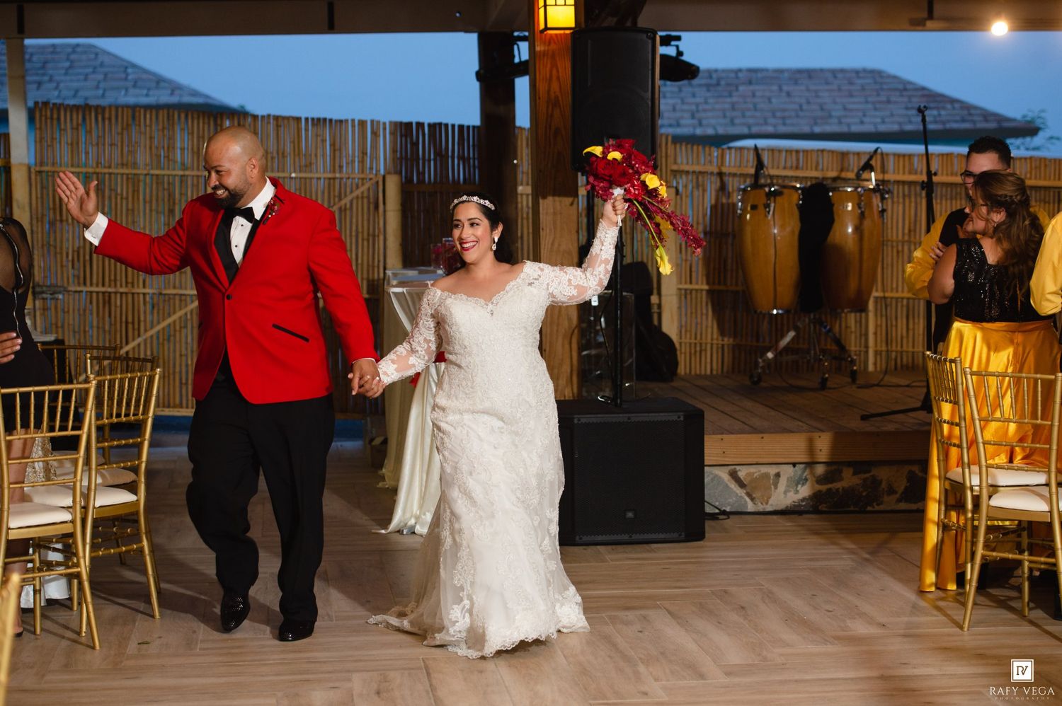 Bride in lace gown and groom in red tuxedo make grand entrance at elegant outdoor wedding reception.