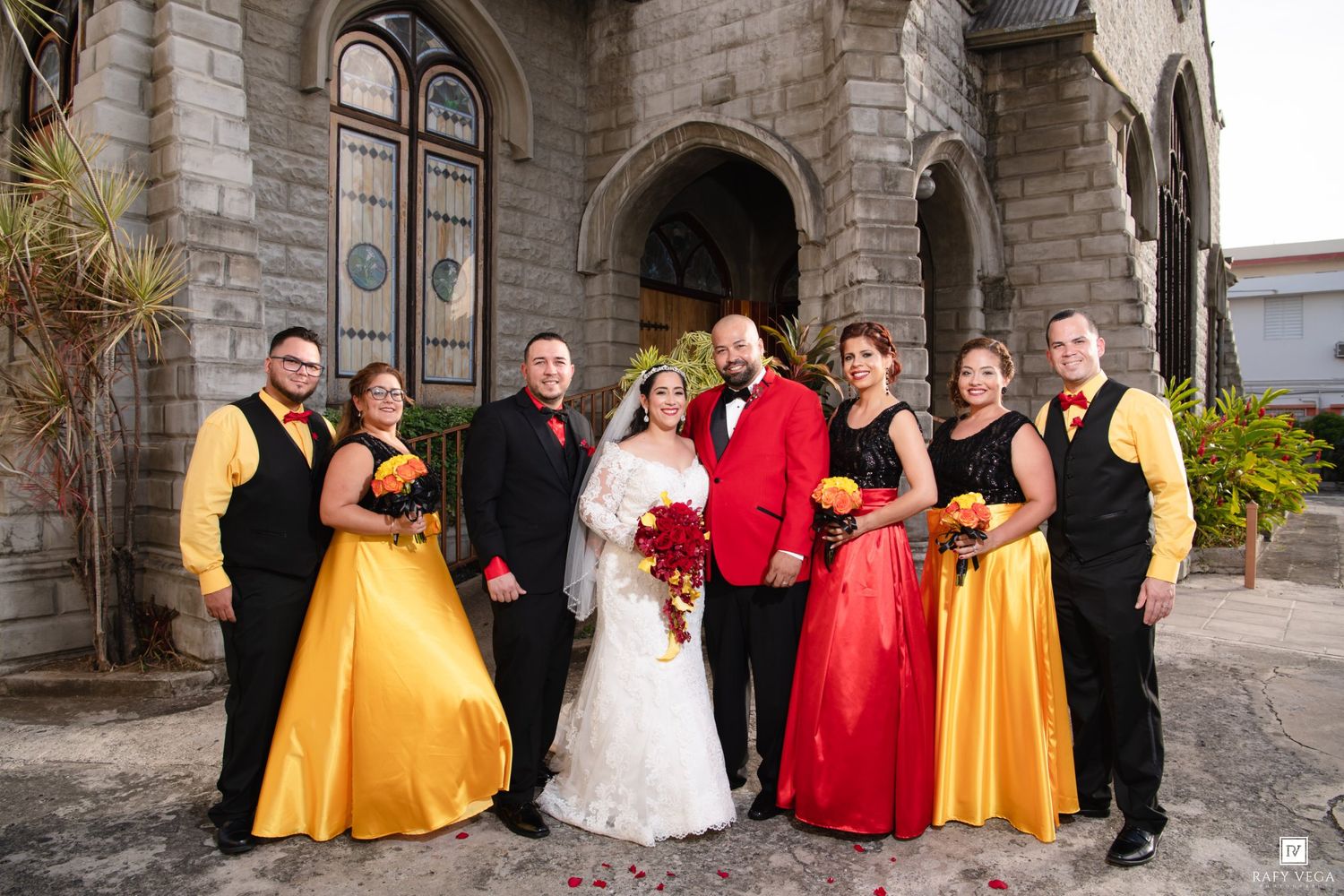 Wedding party poses outside stone church, bride in white, groom in red suit, bridesmaids in yellow and red gowns.