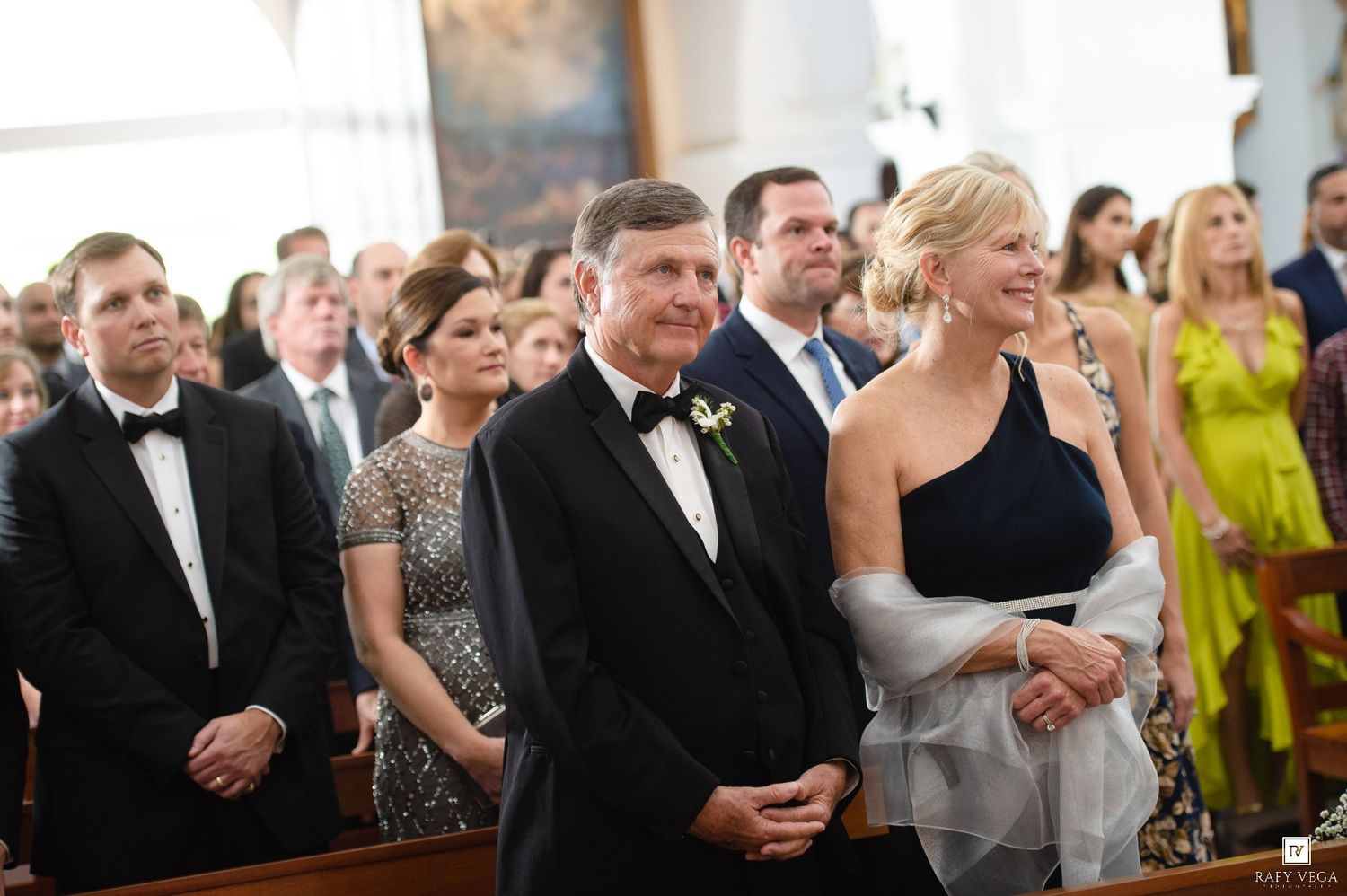 Elegantly dressed wedding guests stand in church pews, smiling during a formal wedding ceremony.