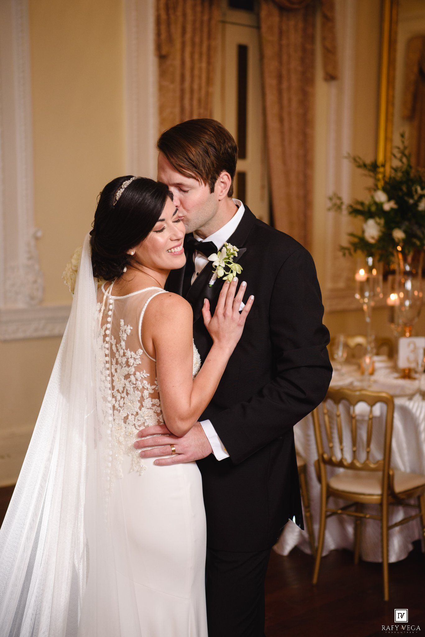 Bride and groom share a tender embrace, with the groom kissing her forehead in an elegant ballroom reception venue.