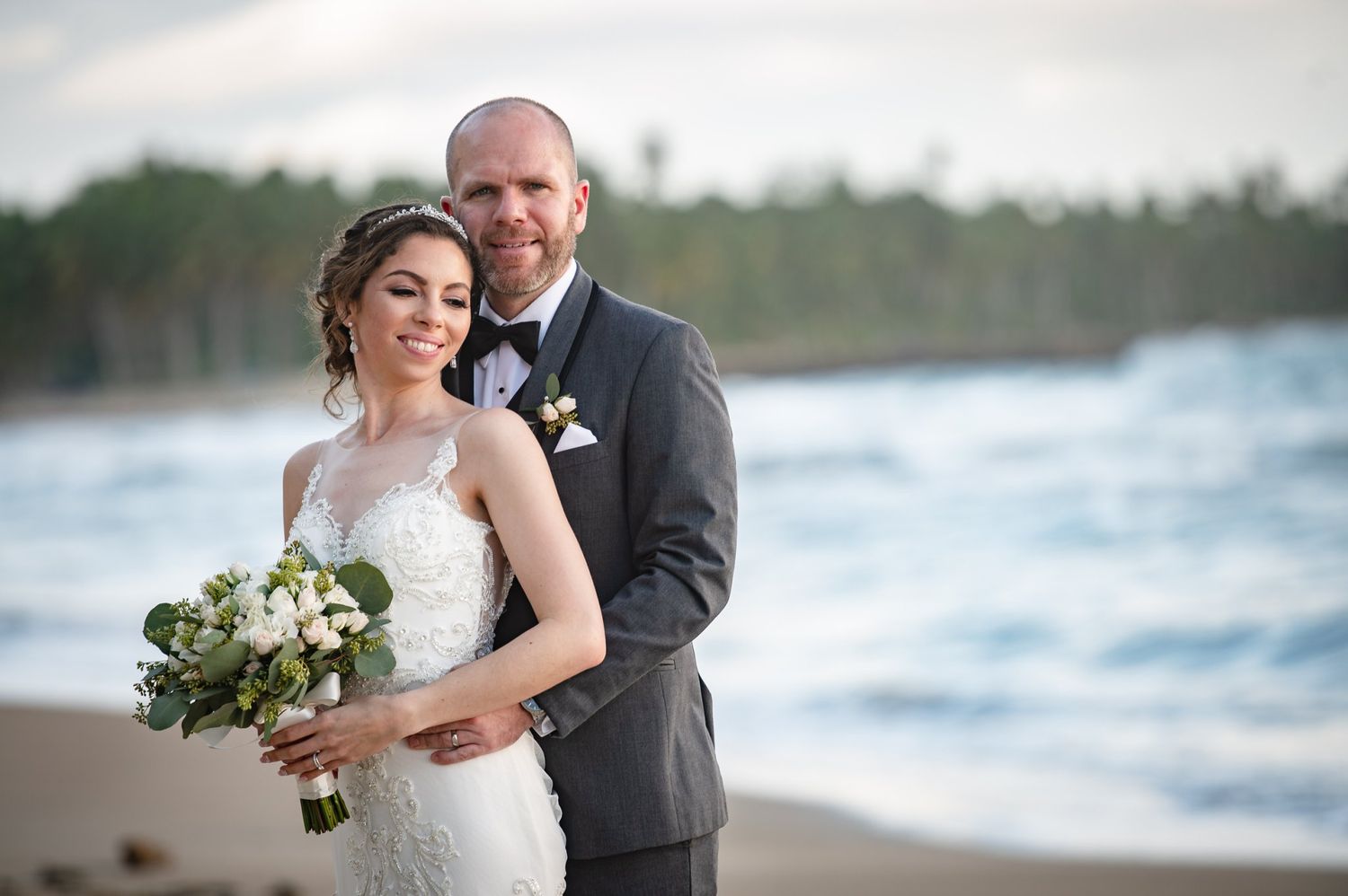 Bride and groom embracing on a beach, bride holding white floral bouquet, groom in gray suit, ocean backdrop.