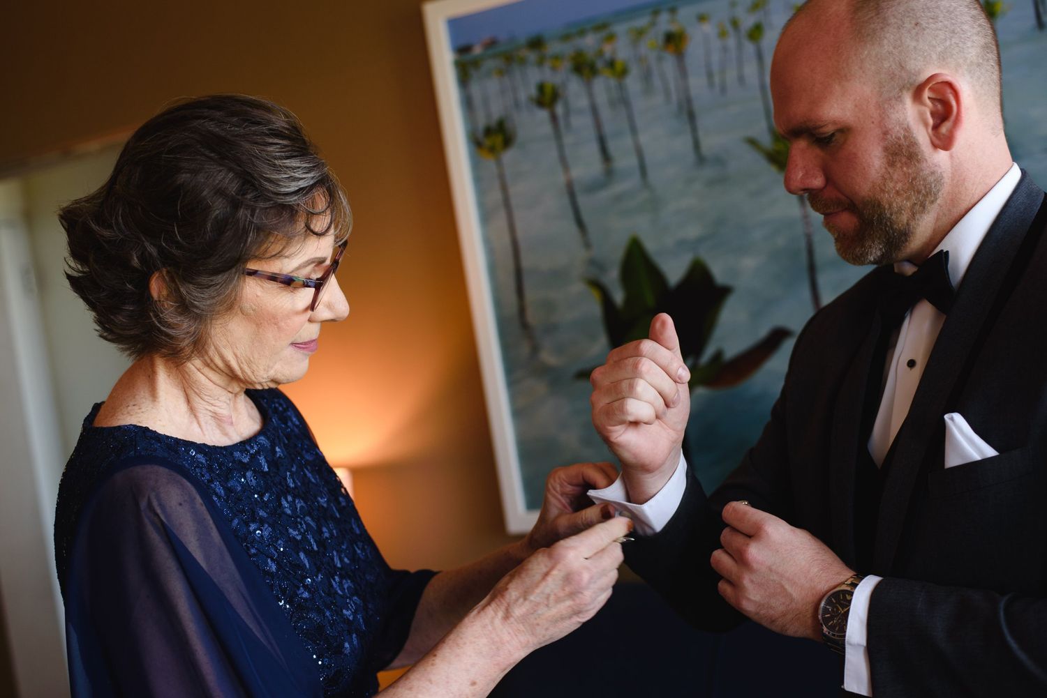 Groom in tuxedo gets cufflinks fastened by older woman in navy dress before wedding ceremony.