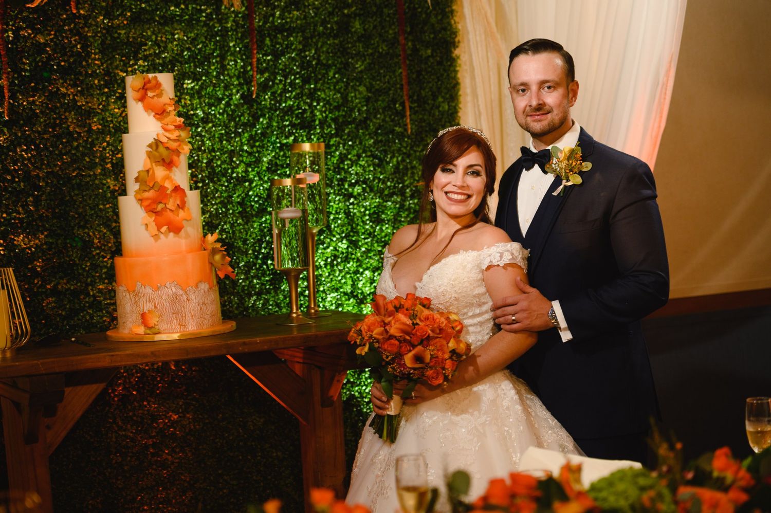 Bride and groom smiling beside a floral wedding cake at an elegant reception with greenery wall backdrop.