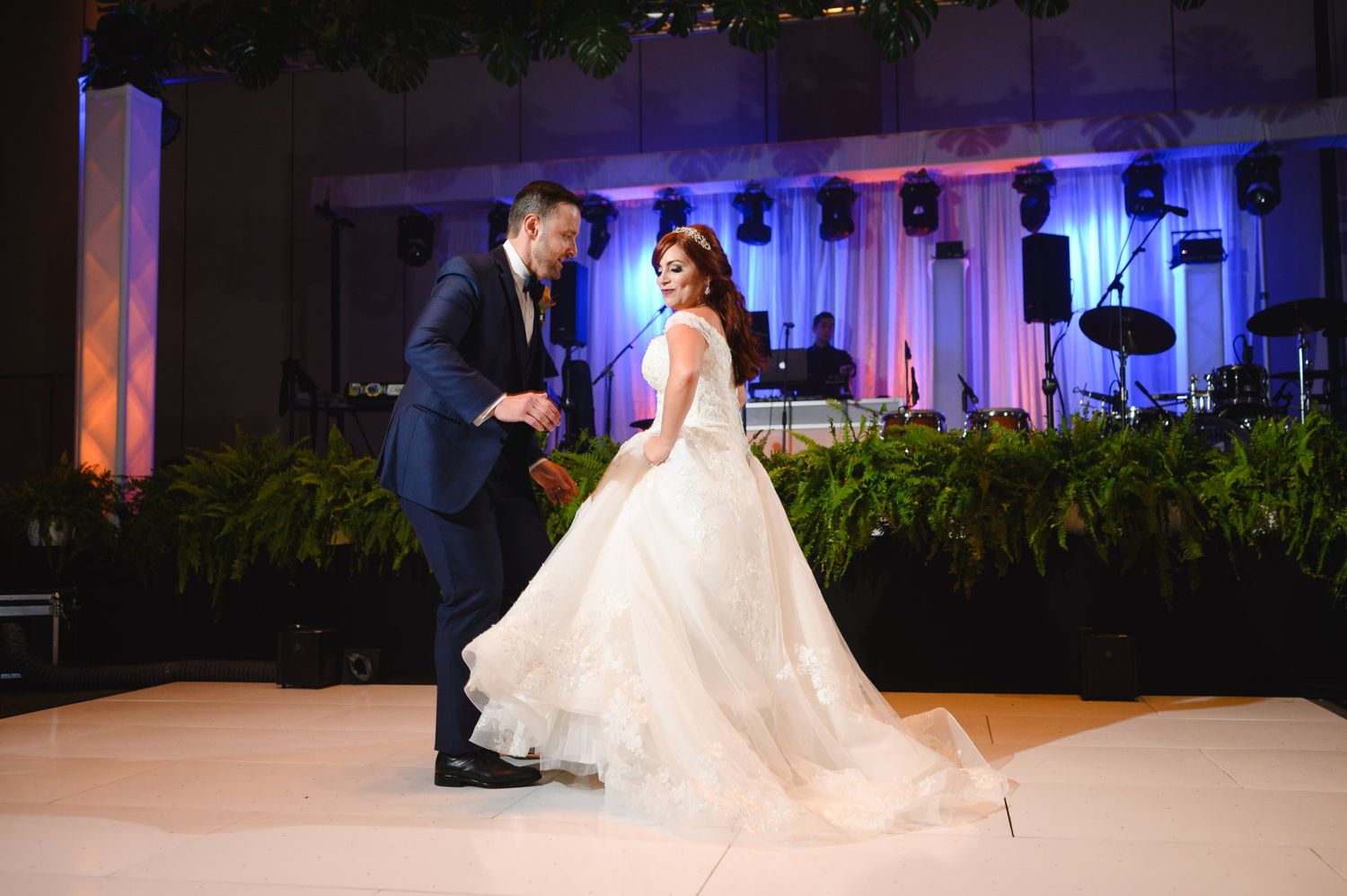Bride and groom share their first dance at elegant wedding reception with blue lighting and floral backdrop.