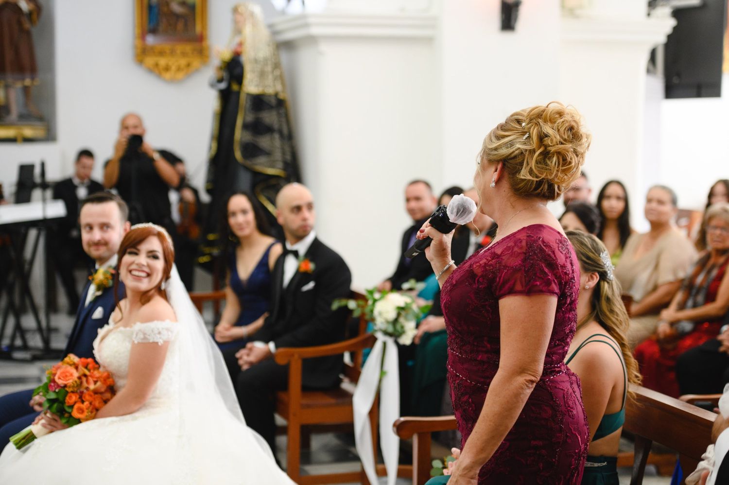 Woman in burgundy dress reads speech at elegant church wedding ceremony with smiling bride in white gown holding orange bouquet.