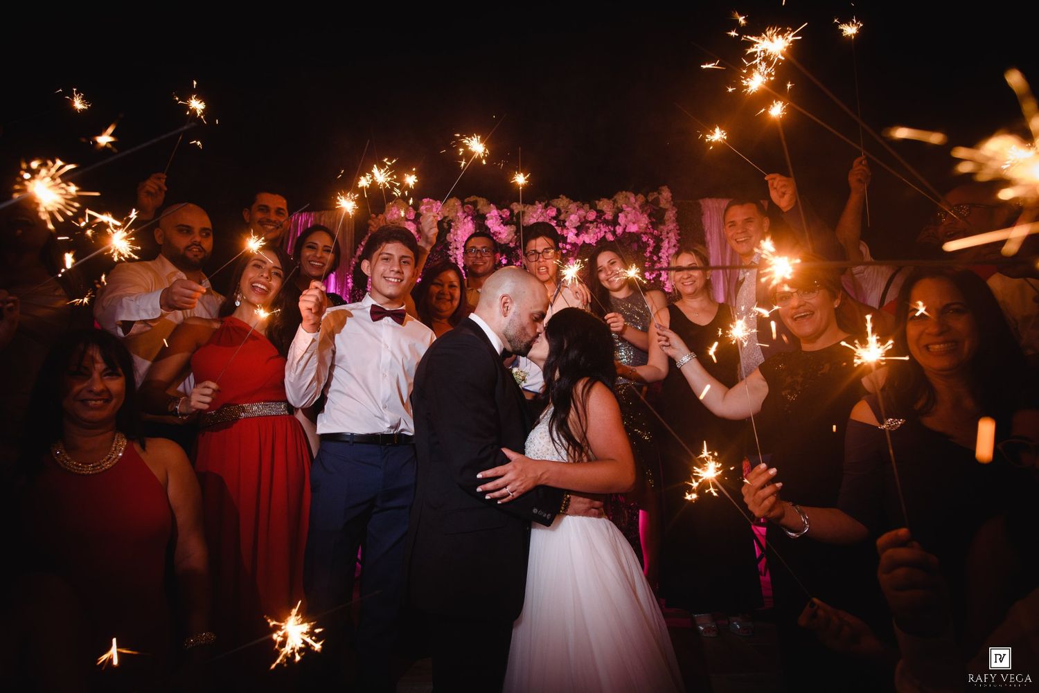 Bride and groom share a romantic kiss surrounded by wedding guests holding sparklers at a nighttime reception.