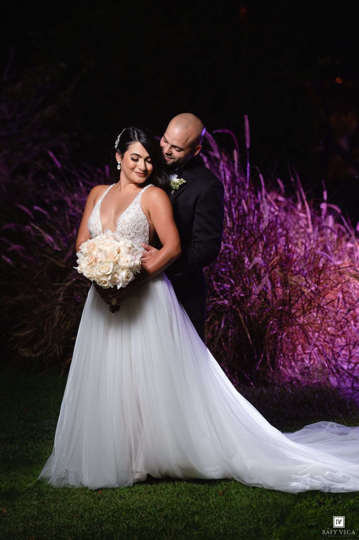 Wedding couple poses romantically at night with purple-lit ornamental grasses backdrop, bride holding white bouquet.