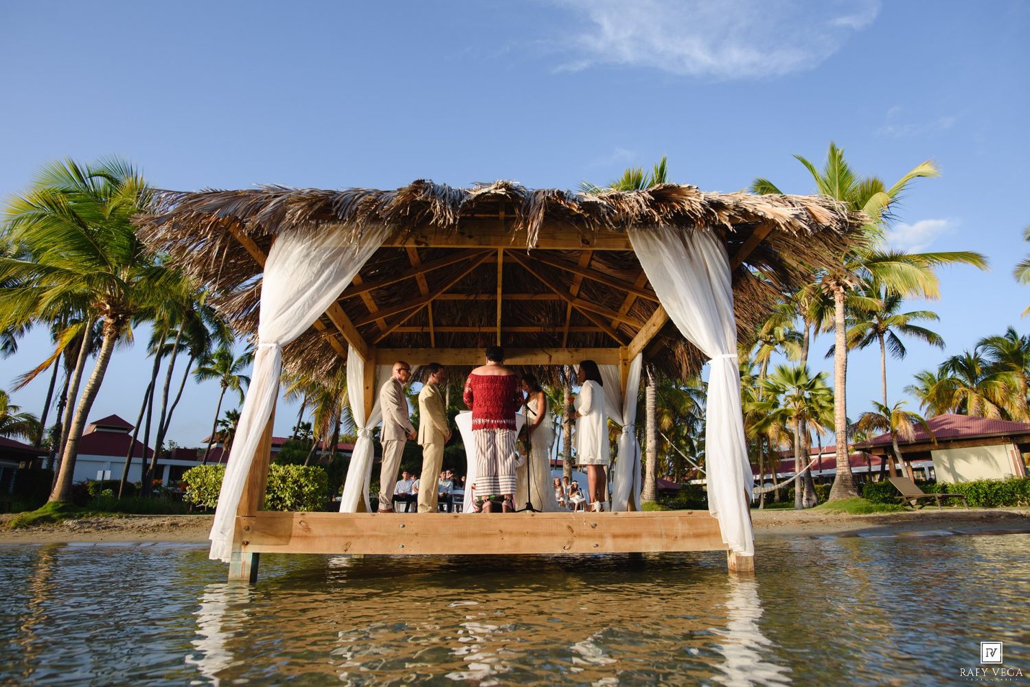 Tropical gazebo wedding ceremony on water surrounded by palm trees under clear blue sky.