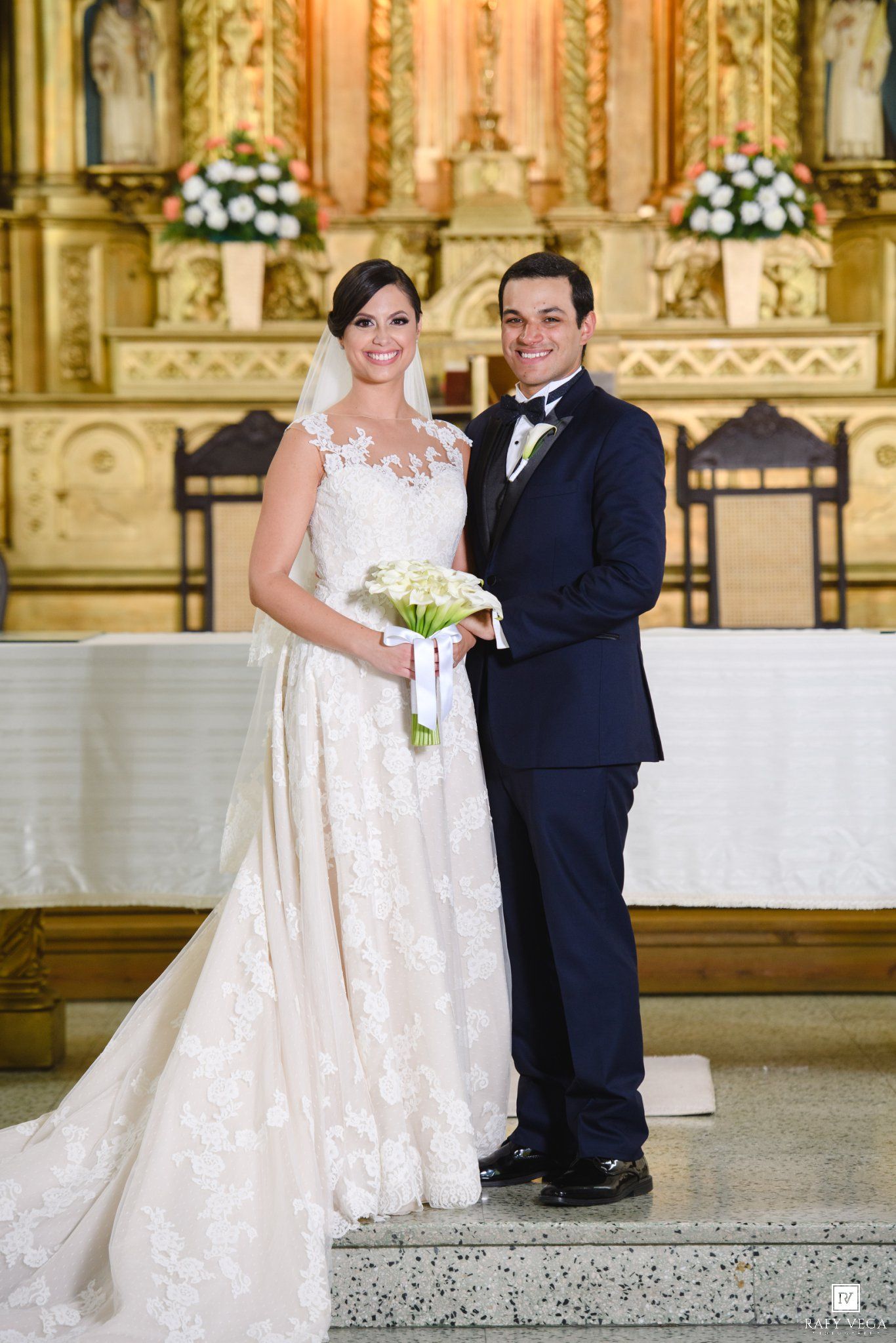 Bride and groom posing together at church altar, bride in lace gown holding white bouquet, groom in navy suit.
