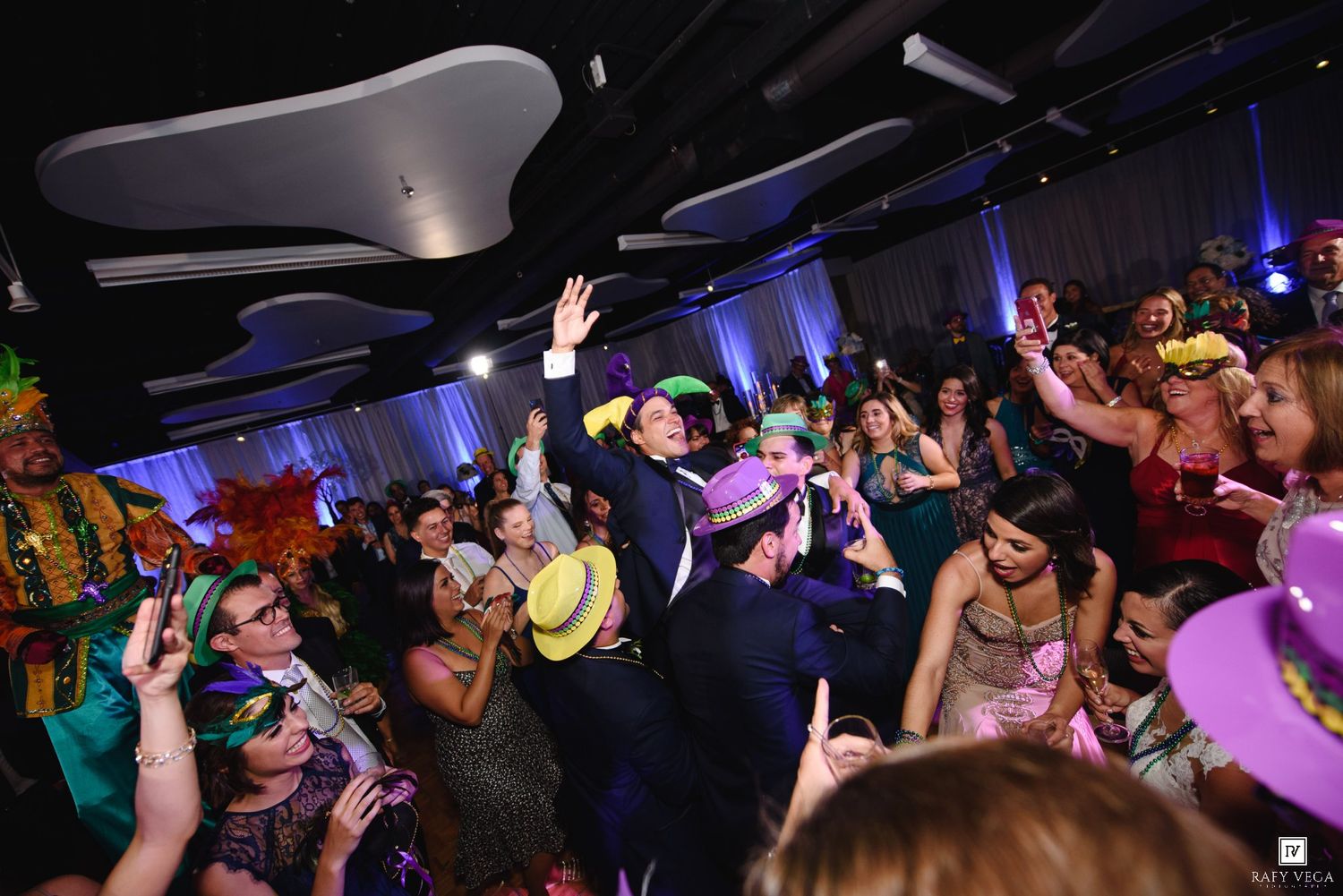 Crowd of party guests in colorful Mardi Gras masks and costumes dancing at a festive masquerade ball event.