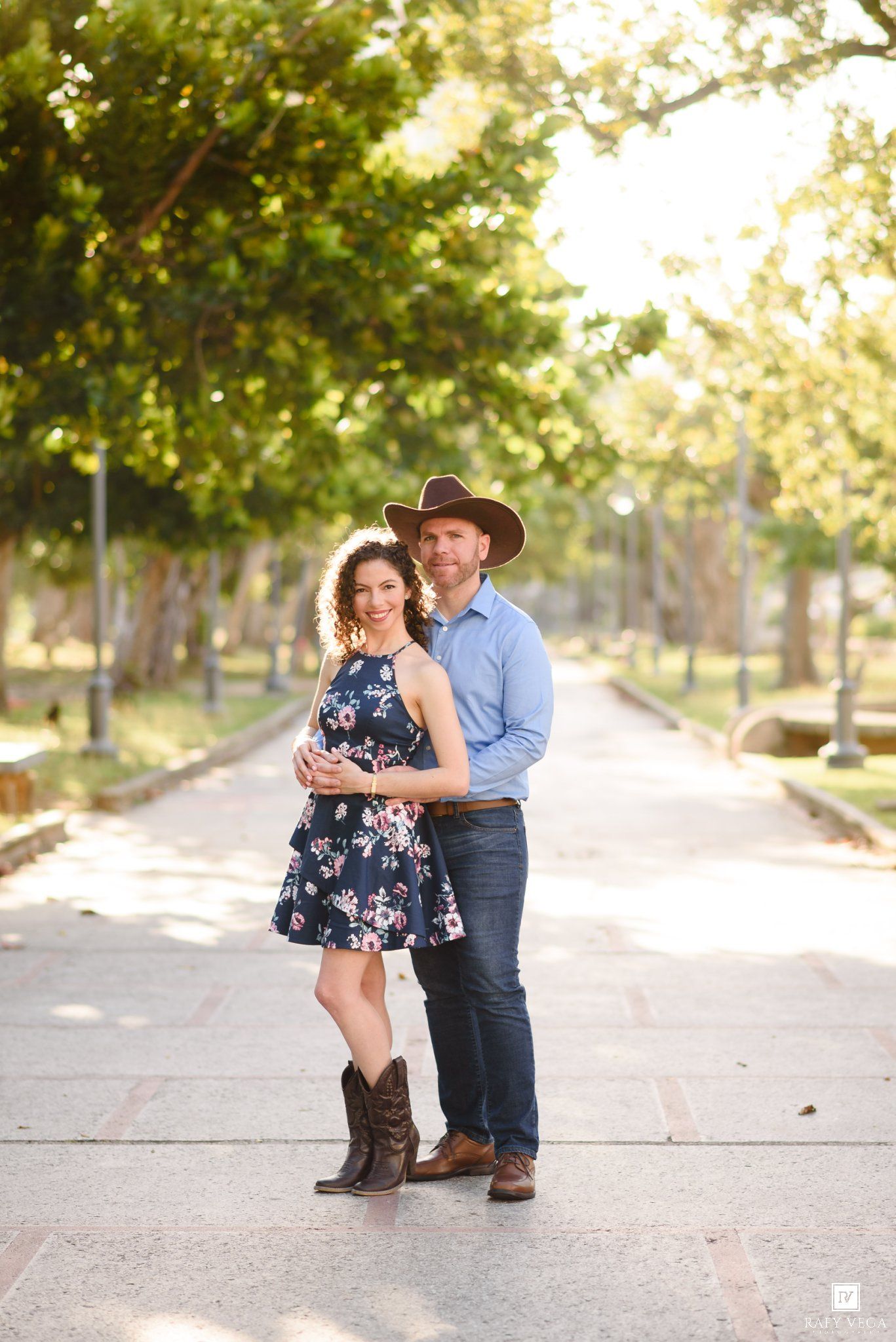 Couple posing on a tree-lined path, woman in floral dress and cowboy boots, man in blue shirt and cowboy hat.