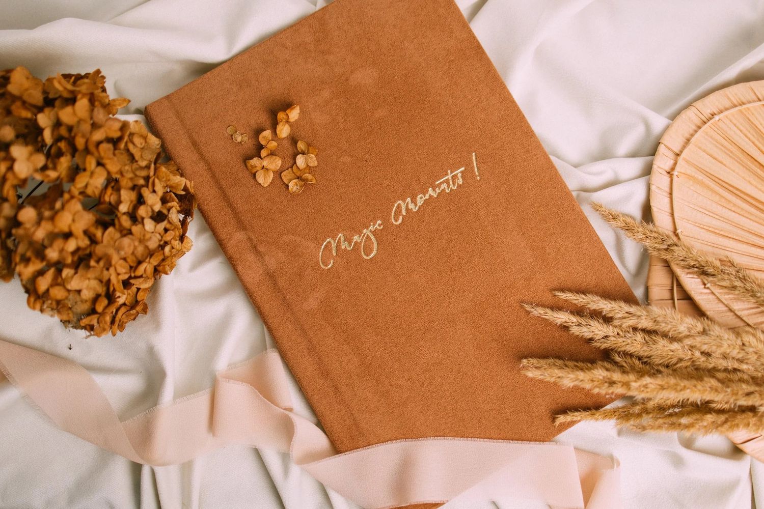Brown leather journal with gold script on white fabric, surrounded by dried wheat stalks and golden nuts.